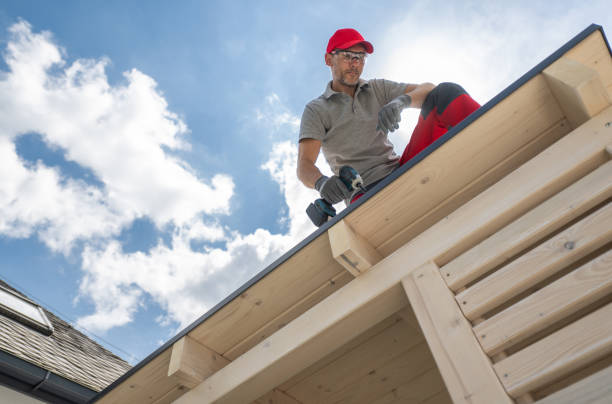 A construction worker in a red hat and safety glasses uses a drill on a new roof. He is working on a sunny day with a blue sky and white clouds. The roof is made of wood and is part of a new construction project.
